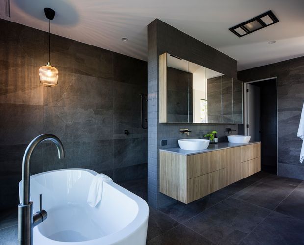 A contemporary bathroom with a freestanding white bathtub, dark large-format tiled walls and floor, a double vanity with wood-grain cabinetry, vessel basins, and mirrored storage. Black fixtures, a sculptural pendant light, and recessed ceiling lighting create a refined, high-contrast finish.