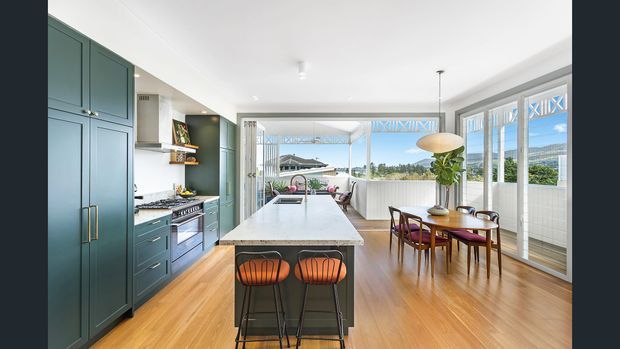 Bright open-plan kitchen and dining area with deep teal built-in cabinetry, brass-toned handles, a large pale stone island with two stools, and warm timber flooring. A dining table sits beside expansive glazed windows and doors opening to a covered outdoor area, with views of greenery and hills beyond. The space combines clean contemporary joinery with mid-century-inspired dining furniture and pendant lighting.