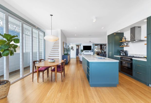 Bright open-plan kitchen and dining area with warm timber flooring, teal-blue island and cabinetry, white walls and ceiling, and large glazed windows with decorative security screens. A round timber dining table sits beneath a pendant light, while the kitchen includes a stone island benchtop, stainless appliances, open shelving, and dark green cabinetry. The space extends toward a living area at the rear.