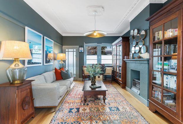 A refined traditional living room with deep blue-grey walls, ornate white ceiling cornices, timber built-in bookcases, a fireplace, upholstered sofas, patterned area rug, and a central coffee table. Decorative mirrors, framed artwork, lamps, and classic furnishings create a warm, layered, library-like atmosphere.