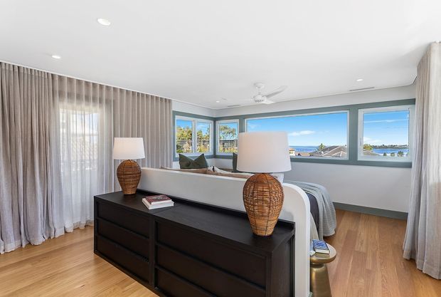Bright coastal bedroom viewed from behind a dark timber dresser, with a bed facing wide blue-grey framed windows overlooking a waterfront or coastal landscape. Floor-to-ceiling sheer and taupe curtains, light timber flooring, bedside lamps, and a ceiling fan create a relaxed, airy interior.