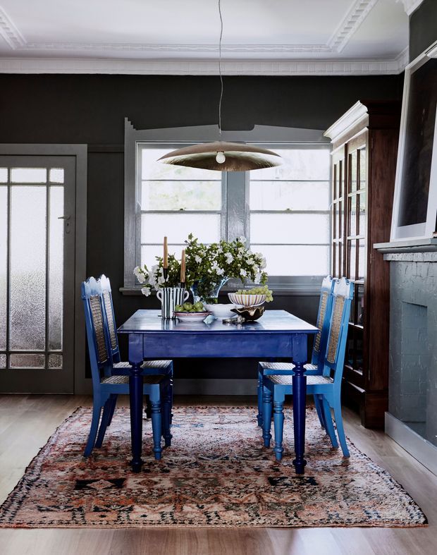 A richly styled dining room featuring a deep blue rectangular dining table and matching blue chairs on a patterned vintage rug. The room has charcoal walls, ornate white ceiling mouldings, large frosted and clear windows, a dark timber glass-front cabinet, and a painted brick fireplace surround. A sculptural pendant light hangs above the table, which is arranged with flowers and decorative tableware.