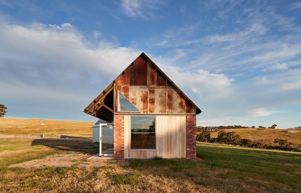 A small rural barn-like building set in an open grassy landscape. The front facade combines weathered corrugated metal, exposed red brick, timber framing and large glazed openings, with a steep gabled roof and a projecting side volume. Warm low-angle sunlight highlights the aged materials against a broad blue sky.