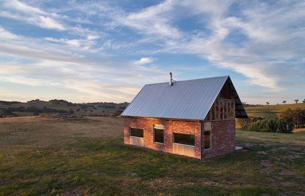 A small standalone rural brick building set in an open rolling landscape. It has a steep pitched gable roof clad in corrugated metal, exposed timber framing in the upper gable, red brick walls, dark window openings, and a simple rectangular form. The scene is photographed at dusk or late afternoon under a partly cloudy sky.