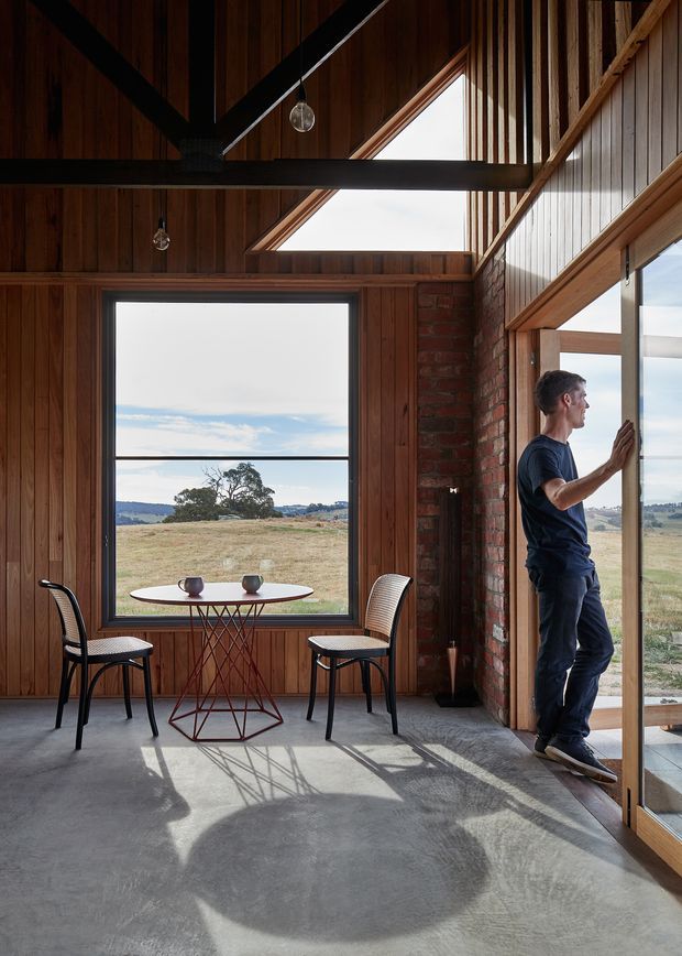 A sunlit interior with a small round dining table and two chairs beside expansive windows overlooking an open rural landscape. The room features warm vertical timber wall and ceiling lining, exposed dark structural beams, a red brick column, polished grey concrete flooring, and large timber-framed glazing. A pitched, barn-like ceiling and clerestory window create a bright, airy atmosphere.