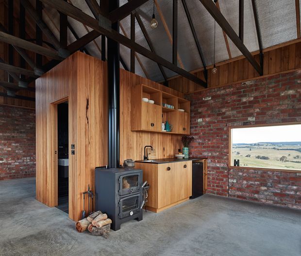 Interior view of a rustic barn-style kitchen and living space with exposed dark timber roof trusses, corrugated metal ceiling panels, red brick walls, polished concrete flooring, and warm timber cabinetry. A compact black wood-burning stove sits in front of the kitchen joinery, with a black rangehood flue, open shelving, sink, and a wide picture window overlooking rolling farmland.