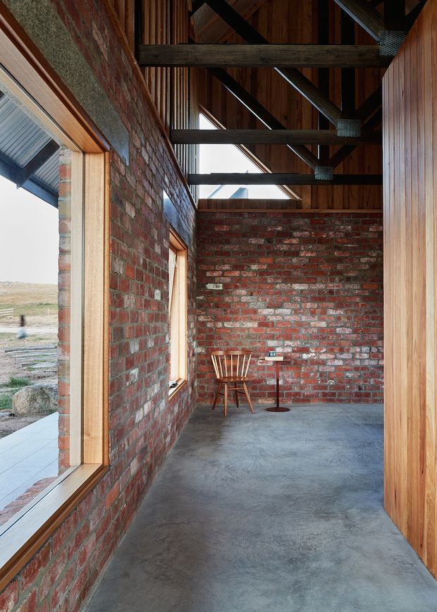 A narrow, rustic-modern interior passage or living nook with exposed red brick walls, polished concrete flooring, timber-framed glazing, and warm vertical timber wall lining. Heavy dark timber roof beams and exposed rafters span overhead, with linear skylights bringing in daylight. A small timber chair and slim round table sit at the far end, while an expansive rural landscape is visible through the windows.