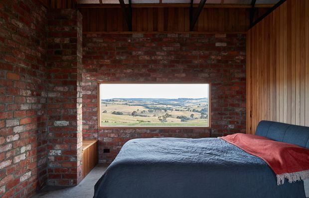 A compact rustic bedroom with exposed red brick walls, dark timber-lined ceiling and vertical timber wall panelling. A bed with dark blue bedding and a red throw dominates the room, while a wide horizontal window frames an expansive rural landscape. The room has a warm, textural, moody character with simple built-in timber joinery.