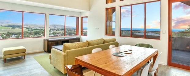 Bright open-plan living and dining area with expansive timber-framed glazing overlooking a coastal hillside and ocean view. A large solid timber dining table with white chairs sits in the foreground, while a mustard sofa, low timber cabinetry, and pale flooring create a warm, relaxed interior. A glazed door opens to an outdoor balcony or terrace.