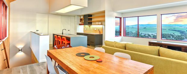 Open-plan dining and living area with a compact contemporary kitchen. A large warm timber dining table occupies the foreground, with white seating and bright orange chairs near the kitchen island. The room features white cabinetry, a pale blue island, timber shelving and accents, tiled flooring, pendant-style ceiling lighting, and expansive windows overlooking a rural landscape.
