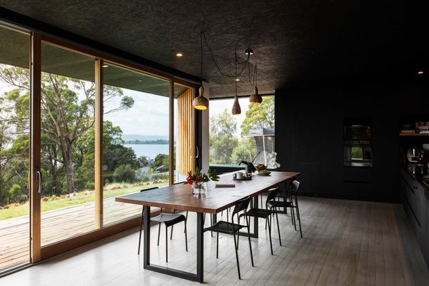 Dark, contemporary dining and kitchen interior with a long timber dining table, black metal-framed chairs, pendant lights, black cabinetry, and expansive timber-framed glazing opening onto a deck. The room has a textured dark ceiling and walls, light timber flooring, and views through the trees toward a body of water.