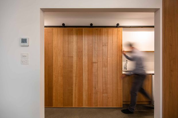 Interior view of a warm timber sliding barn-style door spanning a wide opening, with black exposed roller hardware and a blurred person moving through the adjacent space. White plaster walls, timber detailing, and a grey carpeted floor create a simple, contemporary residential interior.