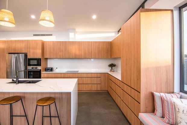 Bright contemporary kitchen with extensive warm timber cabinetry, a large white stone island with integrated sink and seating for two, stainless-steel appliances, white splashback, and a polished concrete floor. A full-height window and built-in timber joinery bring natural light into the space, with woven pendant lights and a small upholstered seating area adding warmth.