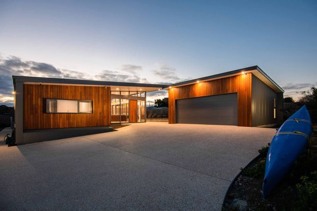 Twilight exterior view of a contemporary low-profile property comprising a timber-clad living wing connected by a glazed entry link to a separate gable-roof garage or outbuilding. The broad concrete driveway dominates the foreground, with warm exterior lighting illuminating the timber facades and large garage door. A blue kayak is visible at the right edge.