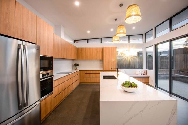 Bright contemporary kitchen with extensive warm timber cabinetry, integrated stainless-steel appliances, a large white stone island with waterfall edge, and black-framed floor-to-ceiling glazing. Pendant lights hang above the island, while clerestory windows bring in additional daylight.