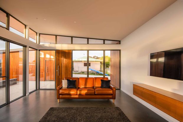 A bright, modern living room with a warm cognac leather sofa facing a wall-mounted television and low built-in timber media console. Full-height black-framed glazing and clerestory windows provide abundant daylight, while timber wall and door panels add warmth. The room has a polished dark floor, white walls and ceiling, and a large dark rug.