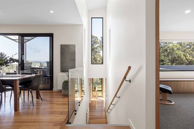 Bright contemporary residential interior centered on a stairwell descending from a timber-floored upper level. The open-plan dining area with dark-framed glazing is visible to the left, while a carpeted room is visible through an opening to the right. White walls, slim vertical windows, timber flooring, a clear glass stair balustrade, and a simple timber handrail create a clean, airy composition.
