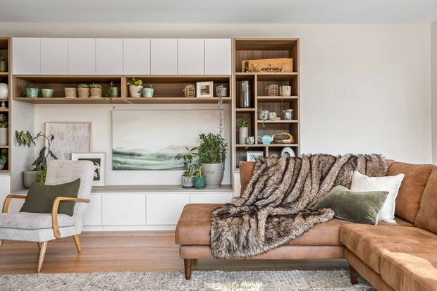 A warm, inviting living room with a built-in white media and display wall, open timber shelving, decorative objects, artwork, indoor plants, and a large tan L-shaped sofa. A textured faux-fur throw and cushions add softness, while a timber-framed armchair and timber flooring complete the space.