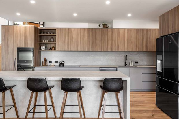 A contemporary kitchen with a long timber-grain wall of overhead cabinetry, integrated appliances, grey lower cabinets, and a large pale stone island with a waterfall-style end. Four dark upholstered timber bar stools line the island. Recessed ceiling lights, open shelving, stainless appliances, and a black refrigerator complete the clean, modern fit-out.