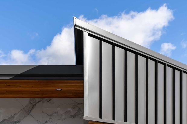 Close-up exterior architectural photograph of a contemporary building against a blue sky. The facade combines tall vertical silver-grey metal panels with dark slender framing, a low dark roof, warm timber soffit, and a marble-look wall surface. The composition emphasizes the crisp geometric roofline and vertical cladding.