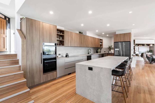 Bright, open-plan contemporary kitchen with extensive timber cabinetry, integrated black appliances, a large pale stone island with bar seating, and warm timber flooring. The kitchen opens directly into a dining and living area, with a stairway and entry door visible at left. Recessed ceiling lighting and clean-lined joinery create a spacious, refined family home interior.