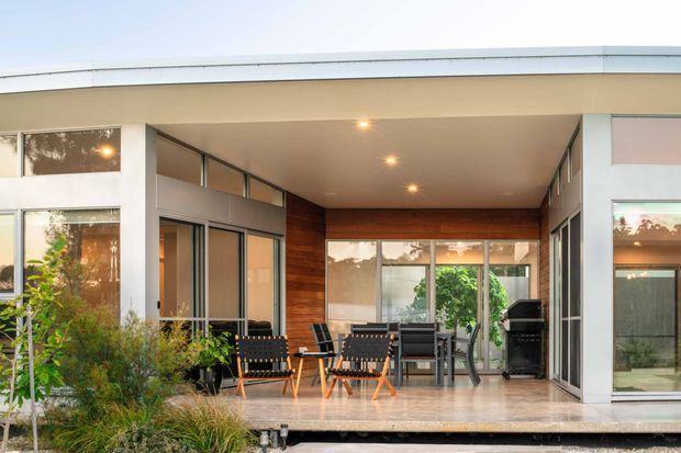 A covered outdoor living terrace beside a modern single-storey home, with broad floor-to-ceiling glazing, slim metal-framed doors and windows, warm timber wall cladding, a pale tiled or stone-like floor, outdoor lounge furniture, and a barbecue. Built-in recessed ceiling lights create a welcoming evening atmosphere, while native-style planting borders the deck.