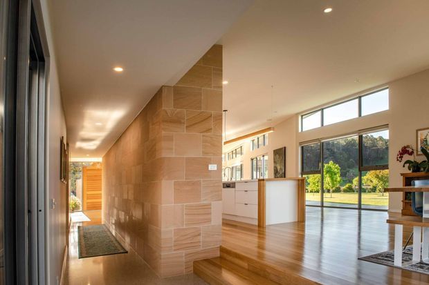 A bright contemporary open-plan home interior viewed from a long entry hall. The hall features a tiled feature wall and polished light flooring, leading toward a timber front door. Beyond the partial partition, a spacious living area has warm timber floors, large floor-to-ceiling windows, a white kitchen island or joinery, and views to a landscaped garden. Recessed ceiling lighting and clean-lined finishes create an airy, refined atmosphere.