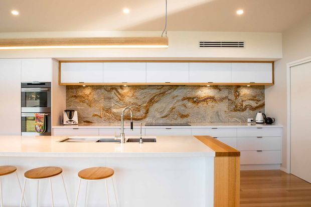 Bright contemporary kitchen with a large white island and three timber-topped stools, extensive white built-in cabinetry, integrated appliances, a timber-framed overhead storage unit, and a dramatic natural stone-look splashback with warm veining. Recessed ceiling lights and under-cabinet lighting create an airy, refined interior.
