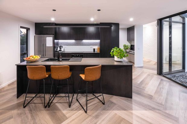 Contemporary open-plan kitchen dominated by a large dark island with three burnt-orange bar stools. The kitchen features dark full-height cabinetry, a stainless-steel refrigerator, integrated appliances, a dark benchtop, and a textured metallic splashback. Pale timber-look flooring is laid in a chevron pattern. Floor-to-ceiling black-framed glazing opens toward a bright adjoining space, with recessed ceiling lighting and indoor greenery adding contrast.