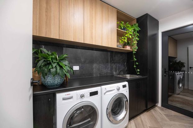 A compact, contemporary laundry room with front-loading white washer and dryer beneath a dark benchtop. The space features dark charcoal tiled splashback and cabinetry, warm timber overhead cupboards, black-framed glazing or a doorway, and indoor plants adding greenery. A mirrored or reflective view reveals additional laundry cabinetry and appliances.
