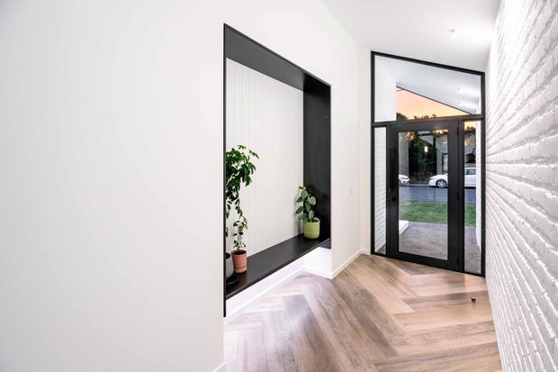 Bright contemporary entry hall with a black-framed glazed front door and transom window, white-painted brick feature wall, pale herringbone timber flooring, and a recessed black-framed display niche containing potted plants. The entrance opens to a small landscaped outdoor area.