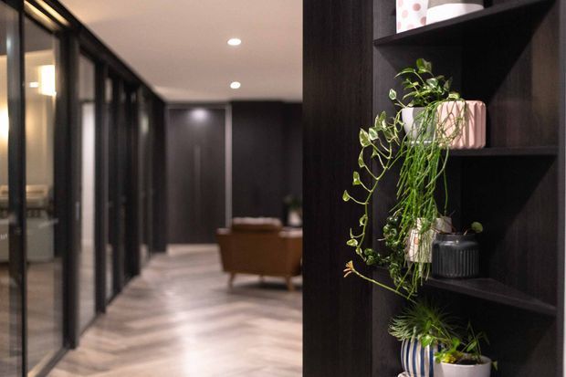 A dark, refined interior hallway with full-height black-framed glazed partitions along one side, pale timber flooring, recessed ceiling lights, and a dark built-in shelving unit displaying potted greenery and decorative objects. A brown armchair and additional seating are visible at the far end.