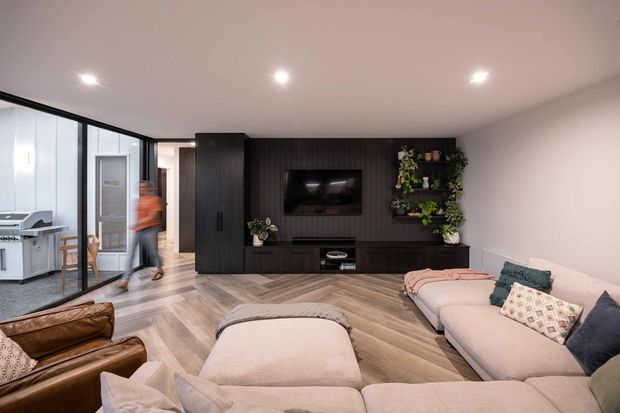 A contemporary open-plan living room with a large sectional sofa and ottoman arranged around a wall-mounted television. A dark, full-height timber-look media wall with integrated cabinetry forms the main feature, alongside a black-framed glazed partition revealing an adjacent workspace or utility area. The room has warm timber flooring, white walls and ceiling, recessed downlights, and a neutral upholstered seating arrangement with dark and patterned cushions.