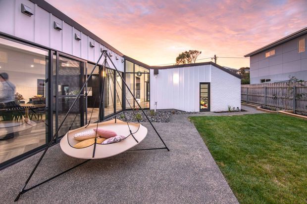 A contemporary single-storey courtyard-style residential exterior at sunset, with a compact white-clad pavilion, dark-framed floor-to-ceiling glazing, and a paved outdoor area featuring a suspended circular swing seat. A small lawn, timber boundary fencing, gravel landscaping, and adjacent modern structures surround the courtyard.