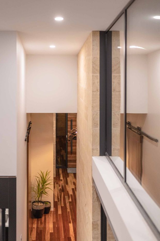 A narrow, modern interior passage viewed beside a large black-framed mirrored or glazed wall. Warm timber flooring leads toward a lower-level area with timber cabinetry and a decorative pendant light. A stone-textured feature wall, white plastered surfaces, recessed downlights, and a simple black handrail create a clean, contemporary circulation space. A potted plant is visible at the base of the passage.