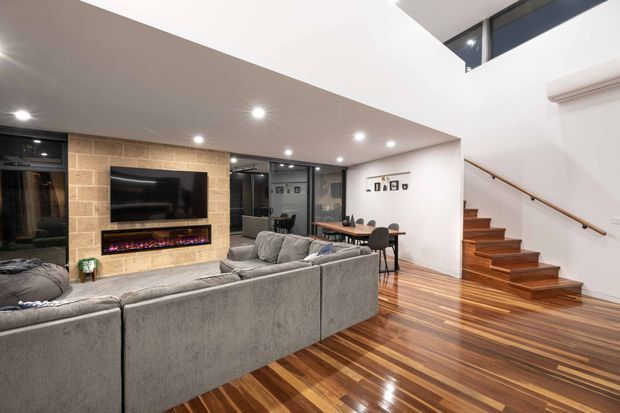 A contemporary open-plan living area with a large grey sectional sofa facing a wall-mounted television and linear electric fireplace set into a tan brick feature wall. The room has glossy striped timber flooring, recessed ceiling lights, extensive dark-framed glazing, and a dining area beside a timber staircase with a simple handrail. A split-system air conditioner is mounted high on the wall.