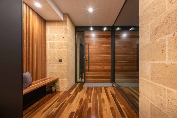 Warm, contemporary entry foyer with a richly patterned timber floor, timber slatted feature wall and built-in bench, textured light stone-block walls, and dark-framed glazed timber entry doors. Recessed ceiling spotlights create a welcoming, refined atmosphere.