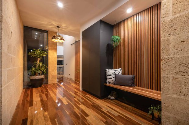 A warm, refined residential hallway or entryway with glossy timber flooring, textured exposed masonry walls, dark built-in cabinetry, and a vertical timber slat feature wall. A built-in timber bench with cushions sits beneath the slatted wall, while recessed ceiling lights and a pendant illuminate the space. A glazed door or opening and indoor planting are visible toward the far end.