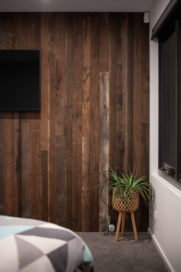 A moody bedroom corner featuring a full-height reclaimed timber feature wall with varied vertical dark wood boards, a wall-mounted black television, dark-framed window, grey carpet, and a potted plant in a woven basket stand. The edge of a bed with geometric black, white, and pale blue bedding is visible in the foreground.