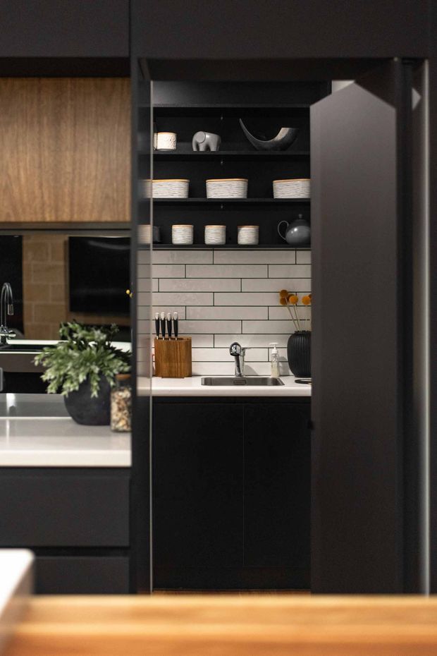A dark, refined kitchen with a compact concealed butler's pantry or preparation nook. The central area features matte-black cabinetry, open black shelving displaying white crockery, a white subway-tile splashback with dark grout, a pale benchtop, stainless-steel sink and tap, and a timber knife block. Dark cabinetry frames the opening, while a warm timber surface appears in the foreground. The adjacent kitchen area is partially visible on the left with additional dark joinery and a pale benchtop.