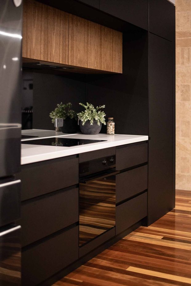 Compact contemporary kitchen with a dramatic black and dark charcoal fit-out, warm vertical-grain timber overhead cabinetry, a white stone-look benchtop, integrated black cooktop and oven, and richly coloured timber flooring. Potted greenery adds a natural accent, while a light exposed-brick wall is visible at the right edge.