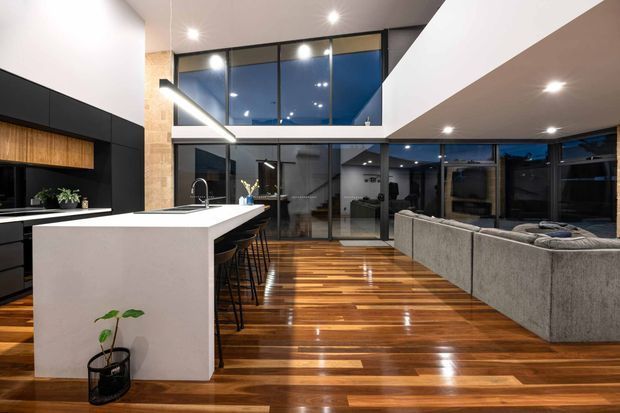 Open-plan contemporary kitchen and living area with a large white island, black and timber cabinetry, integrated sink, bar seating, extensive dark-framed glazing, and a grey sectional sofa. Polished timber flooring and a high ceiling with an upper-level void create a spacious, refined interior.