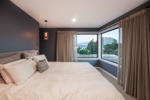 A contemporary bedroom with a large upholstered bed dressed in white bedding, dark blue-grey walls, full-height taupe curtains, carpeted flooring, and a corner of expansive glazing overlooking greenery and distant water or hills. A small pendant light and restrained furnishings create a calm, refined atmosphere.