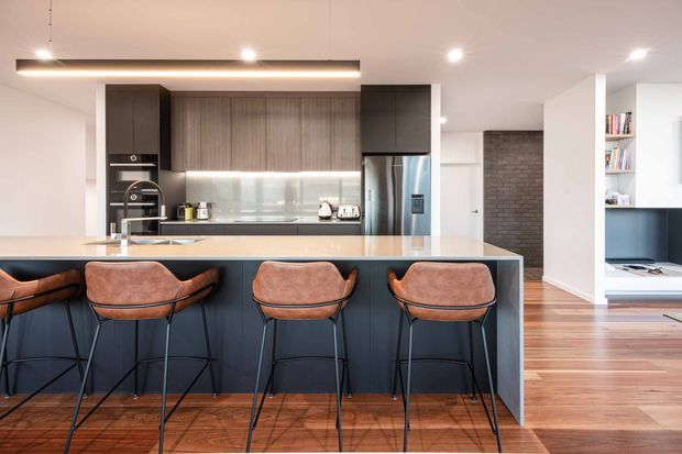 A contemporary open-plan kitchen dominated by a long island with four upholstered bar stools. The kitchen features dark lower cabinetry, warm timber-grain overhead cabinetry, integrated appliances, a pale stone benchtop, and a reflective glass backsplash. Polished timber flooring continues into the adjacent living area, which includes a dark brick feature wall, built-in shelving, and a television. Recessed and linear pendant lighting create a bright, refined interior.