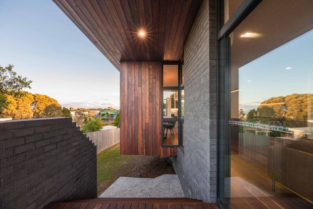 A contemporary residential exterior passage or terrace beside a dark grey masonry wall and large glazed opening. The covered soffit and vertical timber-clad wall create a warm contrast with the cool brickwork, while a narrow stair descends toward a landscaped yard. Reflections in the glazing reveal part of the interior. The elevated view looks across a suburban setting with mature trees.