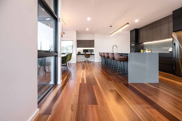 A contemporary open-plan kitchen and dining area with warm timber flooring, dark wood cabinetry, a large grey island with integrated seating, and black-framed glazing along the left side. Recessed and linear pendant lighting illuminate the clean, refined interior.