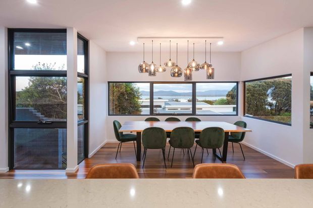 Bright contemporary dining area viewed from behind a pale stone kitchen benchtop. The room features a long timber dining table with green upholstered chairs, polished pendant lights suspended in a linear cluster, white walls and ceiling, dark-framed floor-to-ceiling corner glazing, and broad windows overlooking a coastal or waterside landscape. Timber flooring and extensive glazing create an airy, connected interior.