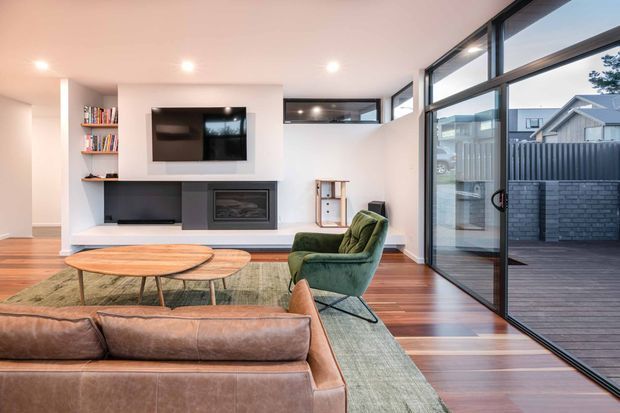 Bright contemporary living room with a low built-in media wall, open shelving, fireplace, green lounge chair, and a large brown leather sofa. Warm timber flooring and expansive black-framed corner glazing open onto a timber deck and fenced exterior courtyard.