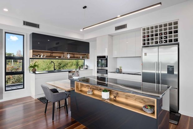 A bright contemporary kitchen with glossy white full-height cabinetry, dark feature cabinetry, stainless-steel appliances, a large island with a timber display niche and stone benchtop, and a built-in wine rack above the refrigerator. A dining table and chairs sit beside the island, with large windows bringing in natural light and views of greenery.