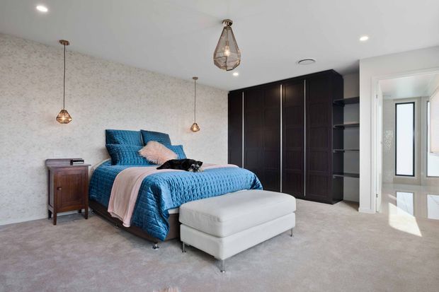 A bright contemporary bedroom with a blue upholstered bed, layered blue and blush bedding, a white upholstered bench, plush carpet, dark full-height built-in wardrobes, and textured patterned wallpaper. Pendant lights and recessed ceiling lighting add warmth, while an open doorway leads to a bright adjacent area.